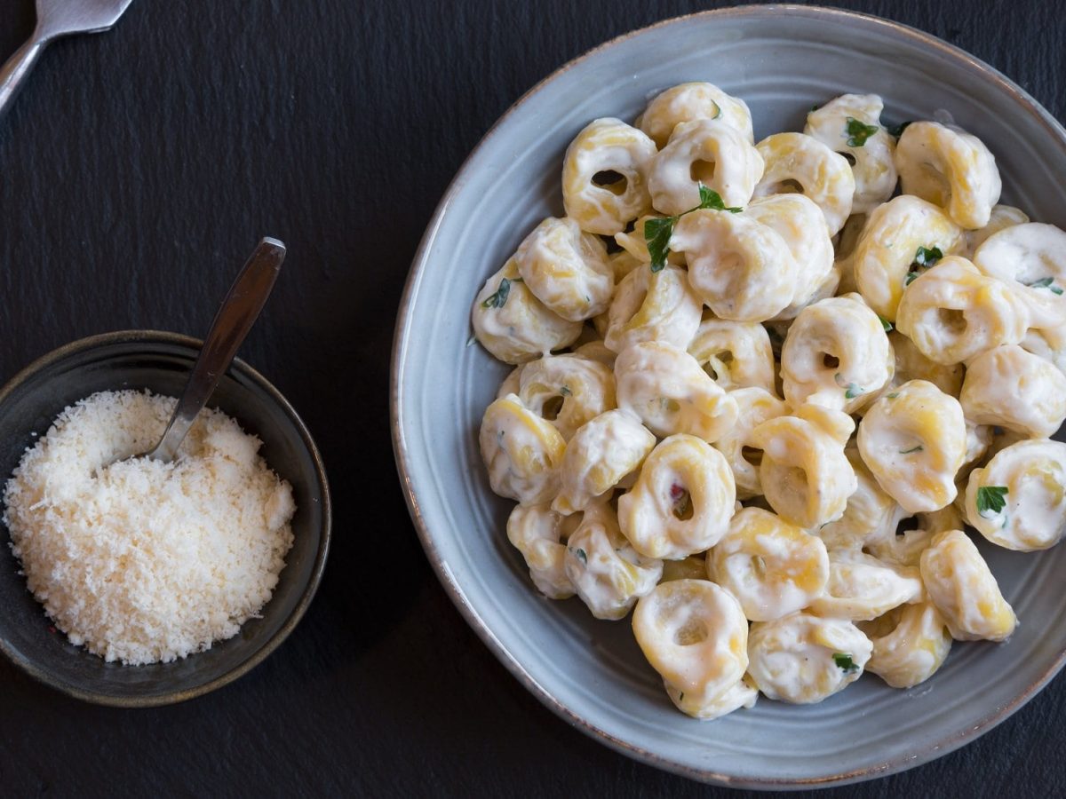 Tortellini in Knoblauch-Sahne-Sauce auf hellem Teller, angerichtet auf dunklem Untergrund. Daneben steht eine Schale mit geriebenem Käse.