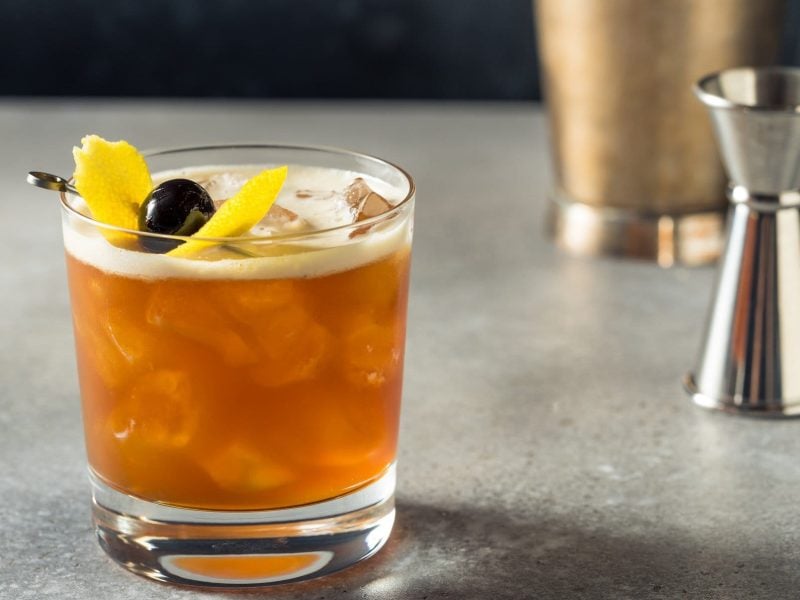 Amaretto Sour mit Zitronenzeste und Cocktailkirsche im Old Fashioned Glas auf grauem Tisch. Im Hintergrund Barmaß und Shaker. Frontalansicht.