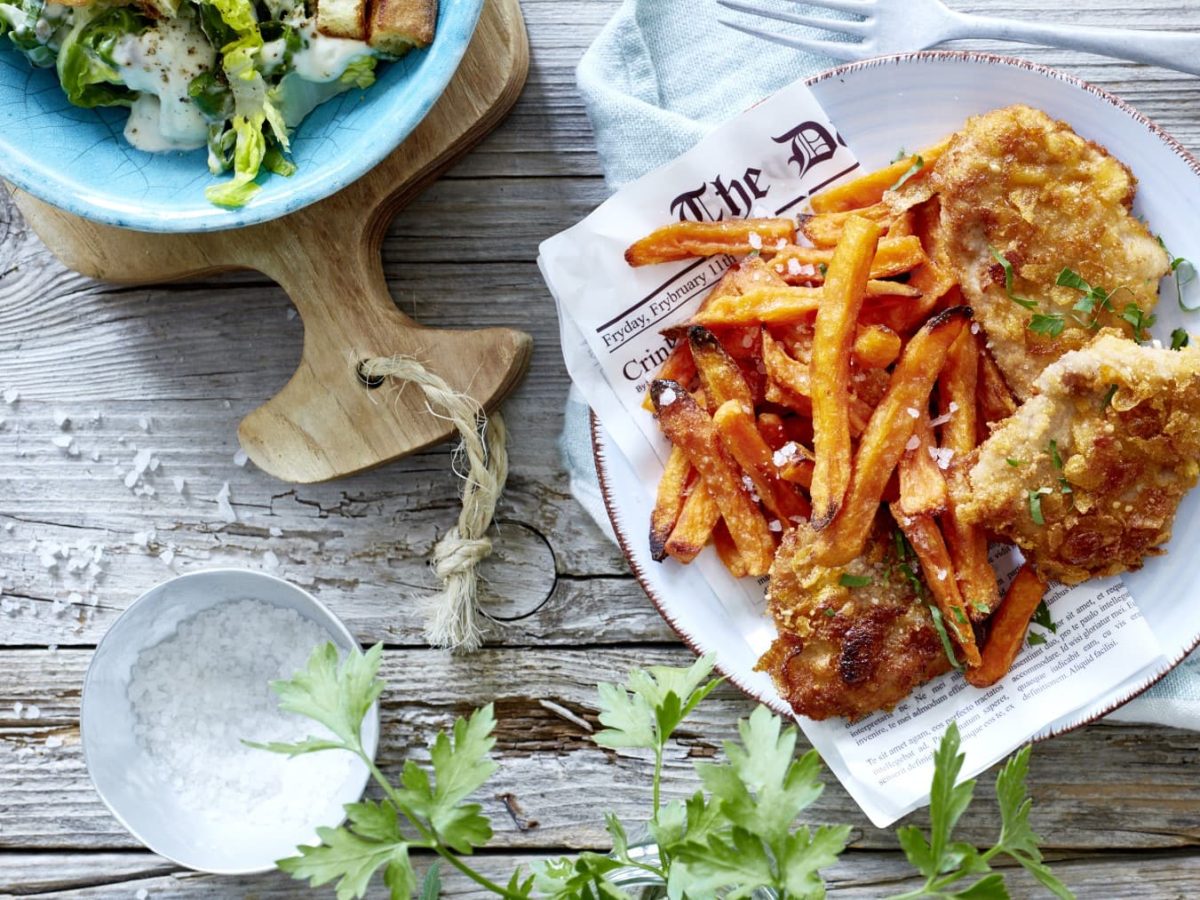 Amerikanische Crisp-Schnitzel mit Süßkartoffel-Pommes auf einem Teller neben einer Schale Salz und einem Teller mit Salat vor Holzhintergrund