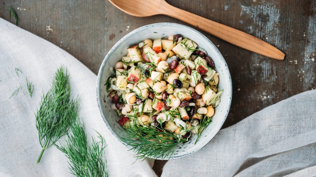 Eine Schale Apfel-Bohnensalat mit Kichererbsen und Dill in der Draufsicht, daneben ein Holzlöffel und frischer Dill.