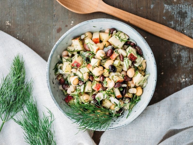 Eine Schale Apfel-Bohnensalat mit Kichererbsen und Dill in der Draufsicht, daneben ein Holzlöffel und frischer Dill.