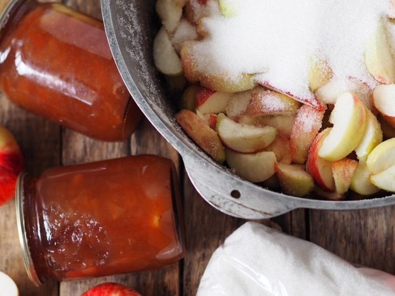 Mehrere Gläser Apfel-Marmelade liegen auf einem Untergrund aus Holz. Neben ihnen liegen Äpfel. Die einen sind ganz, andere sind halbiert. In einer Schale aus Metall liegen Apfelachtel, die mit Zucker bestreut sind.