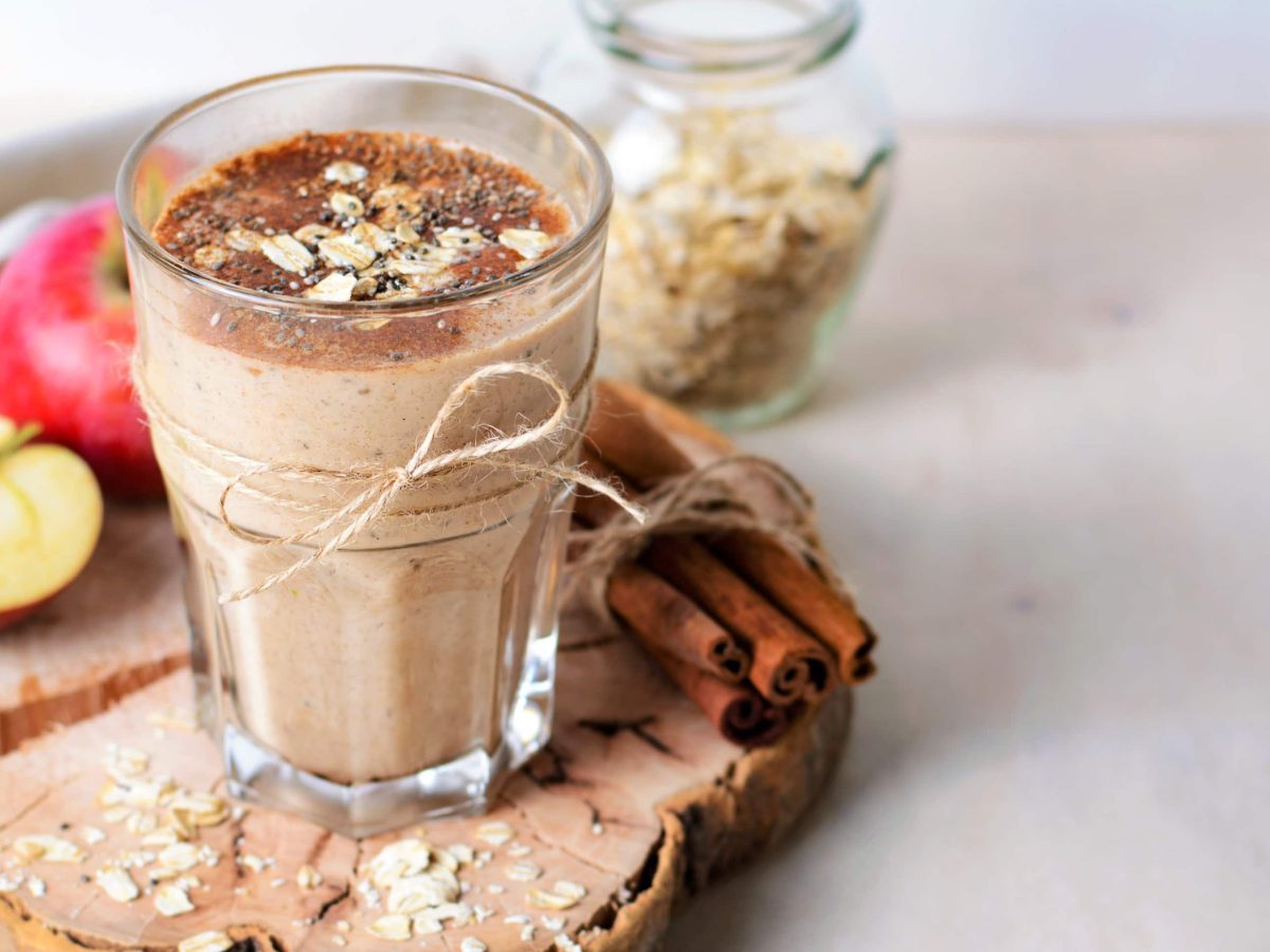 Ein Glas mit dem Apfel-Zimt-Smoothie auf einer Holzscheibe mit frischen Äpfeln und Haferflocken im Hintergrund.