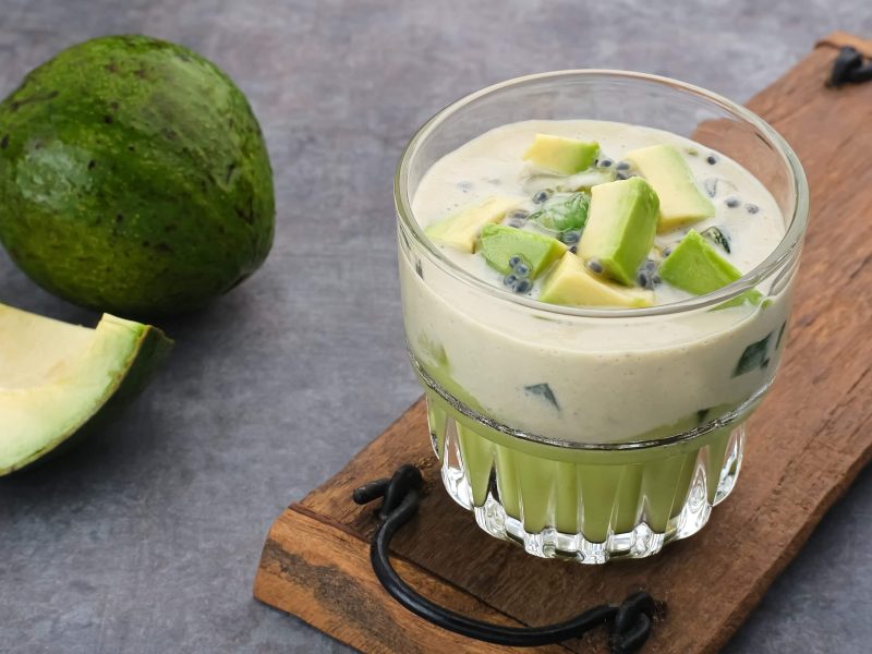 Avocado und Kondensmilch in einem Glas mit Basilikumsamen auf einem Holzbrett und grauem Untergrund. Im Hintergrund eine angeschnittene Avocado.