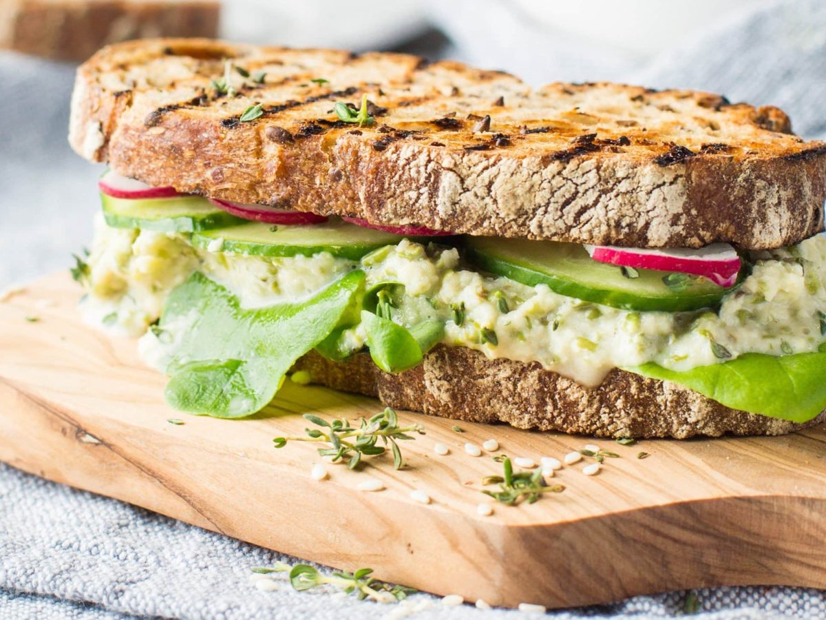 Ein belegtes Sandwich mit Bohnen-Hummus auf einem hellen Holzbrett und einer hellblauen Decke als Hintergrund.