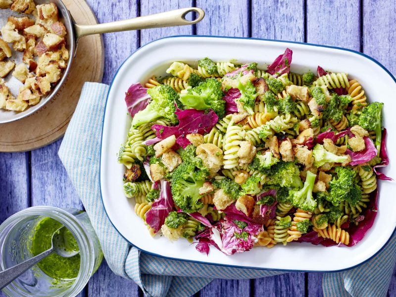 Schüssel mit Broccoli-Nudelsalat neben Schüssel mit Brotbröseln und Glas mit Pesto auf blauen Holzhintergrund