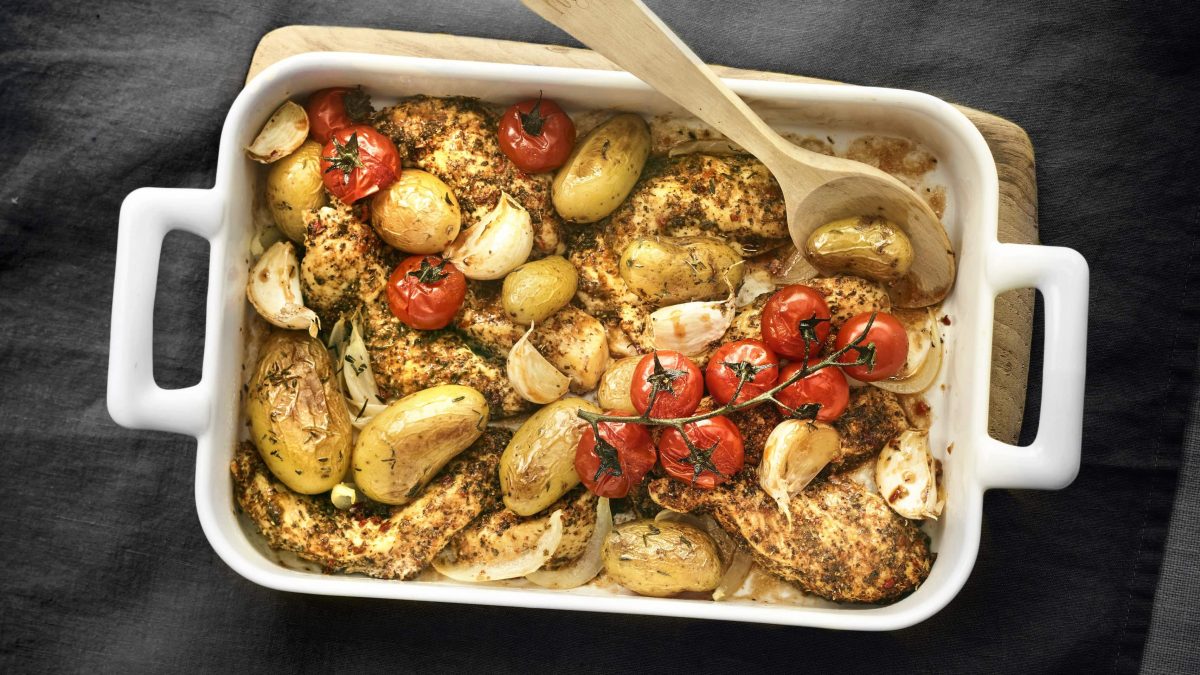 Cajun-Hähnchenbrust mit Bratkartoffeln und Strauchtomaten mit Holzlöffel in einer weißen Ofenform vor dunklem Hintergrund