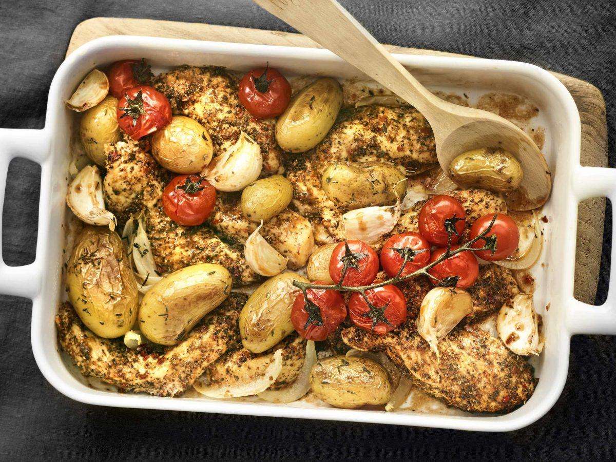 Cajun-Hähnchenbrust mit Bratkartoffeln und Strauchtomaten mit Holzlöffel in einer weißen Ofenform vor dunklem Hintergrund