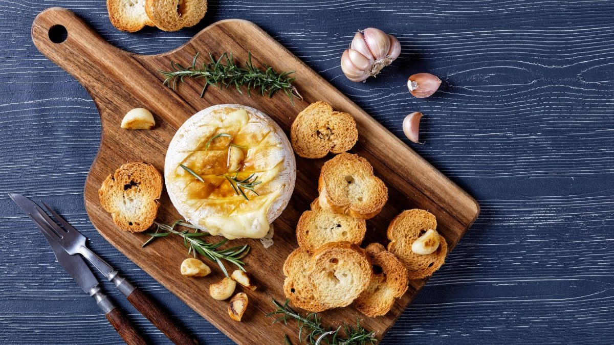 Camembert im Ofen gebacken mit Rosmarinzweigen, Knoblauchzehen und Lorbeerblättern serviert mit Brot auf einem Holzbrett auf dunklem Holzhintergrund, Draufblick, Nahaufnahme