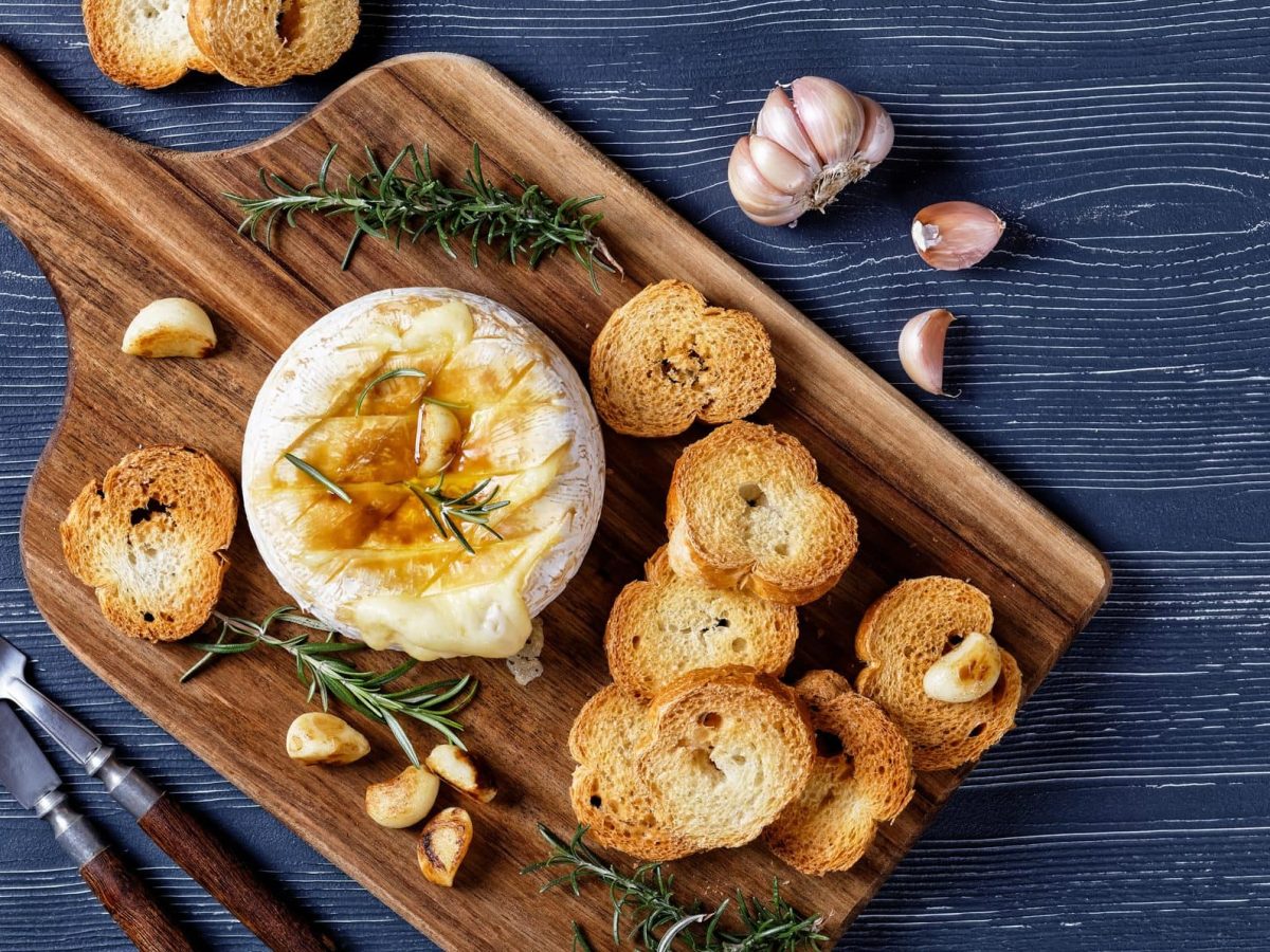 Camembert im Ofen gebacken mit Rosmarinzweigen, Knoblauchzehen und Lorbeerblättern serviert mit Brot auf einem Holzbrett auf dunklem Holzhintergrund, Draufblick, Nahaufnahme