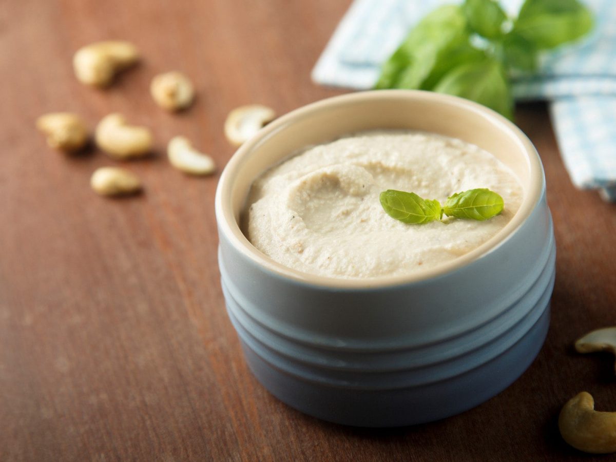 Cashew Creme in blauer Tonschale auf Holztisch. Daneben Cashews, Basilikum und blaues Tuch. Frontalansicht.