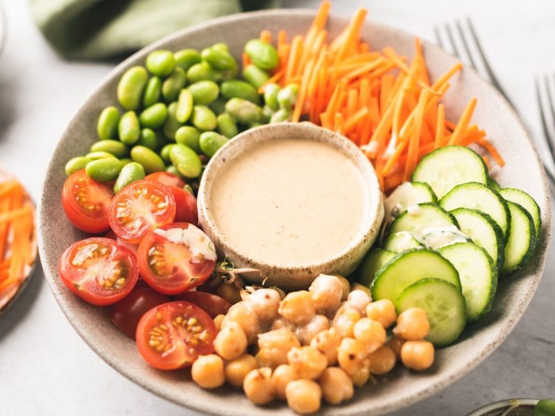 EIn Teller mit klein geschnittenen Karotten, Tomaten, Gurke, Kichererbsen und Edamame, in der Mitte steht ein Schälchen Cashew-Dressing.