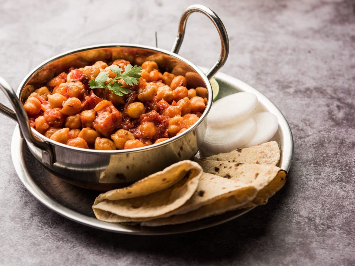 Chana Masala in silberner Schale auf Teller mit Brot auf grauem Untergrund. Draufsicht.