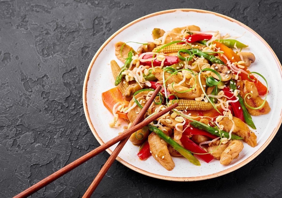 Ein weißer Teller mit dem Chop Suey auf einem schwarzen Untergrund und zwei Essstäbchen am unteren Tellerrand.