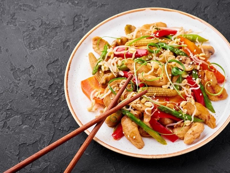 Ein weißer Teller mit dem Chop Suey auf einem schwarzen Untergrund und zwei Essstäbchen am unteren Tellerrand.