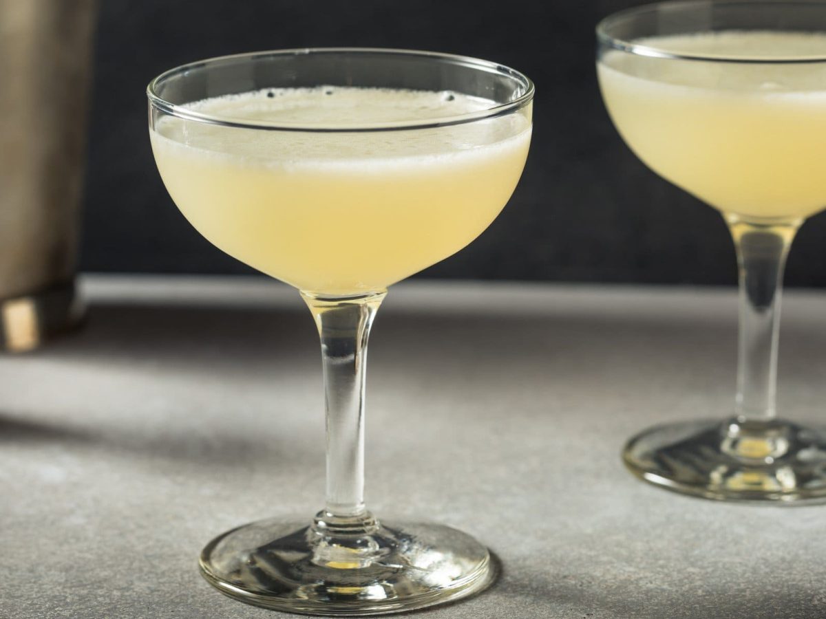 Zwei Corpse Reviver No. 2 auf grauem Tisch vor schwarzem Hintergrund. Dahinter Barmaß und Shaker. Frontalansicht