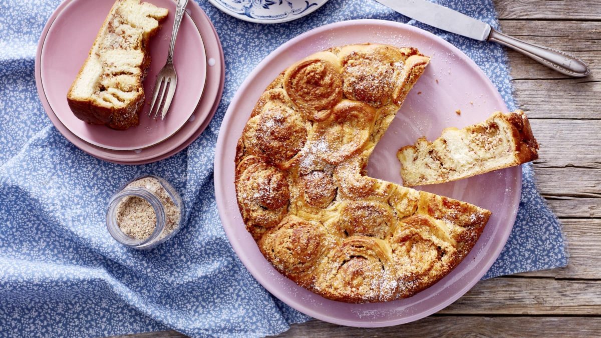 Angeschnittener dänischer Buteerkuchen auf einem rosa Teller, daneben noch ein Teller mit einem Stück Kuchen auf blauer Tischdecke