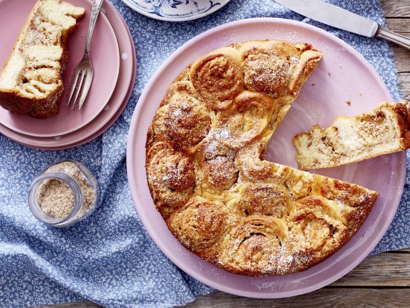 Angeschnittener dänischer Buteerkuchen auf einem rosa Teller, daneben noch ein Teller mit einem Stück Kuchen auf blauer Tischdecke