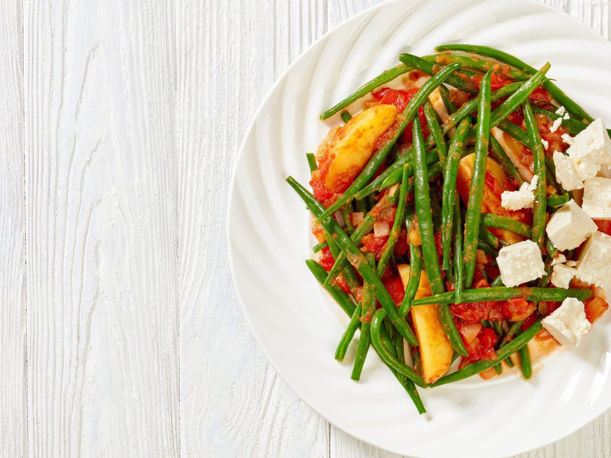 Ein weißer Teller mit grünen Bohnen, gekochten kartoffeln, Tomaten und feta steht auf einem weißen Untergrund. Das gericht heißt Fasolakia lathera.