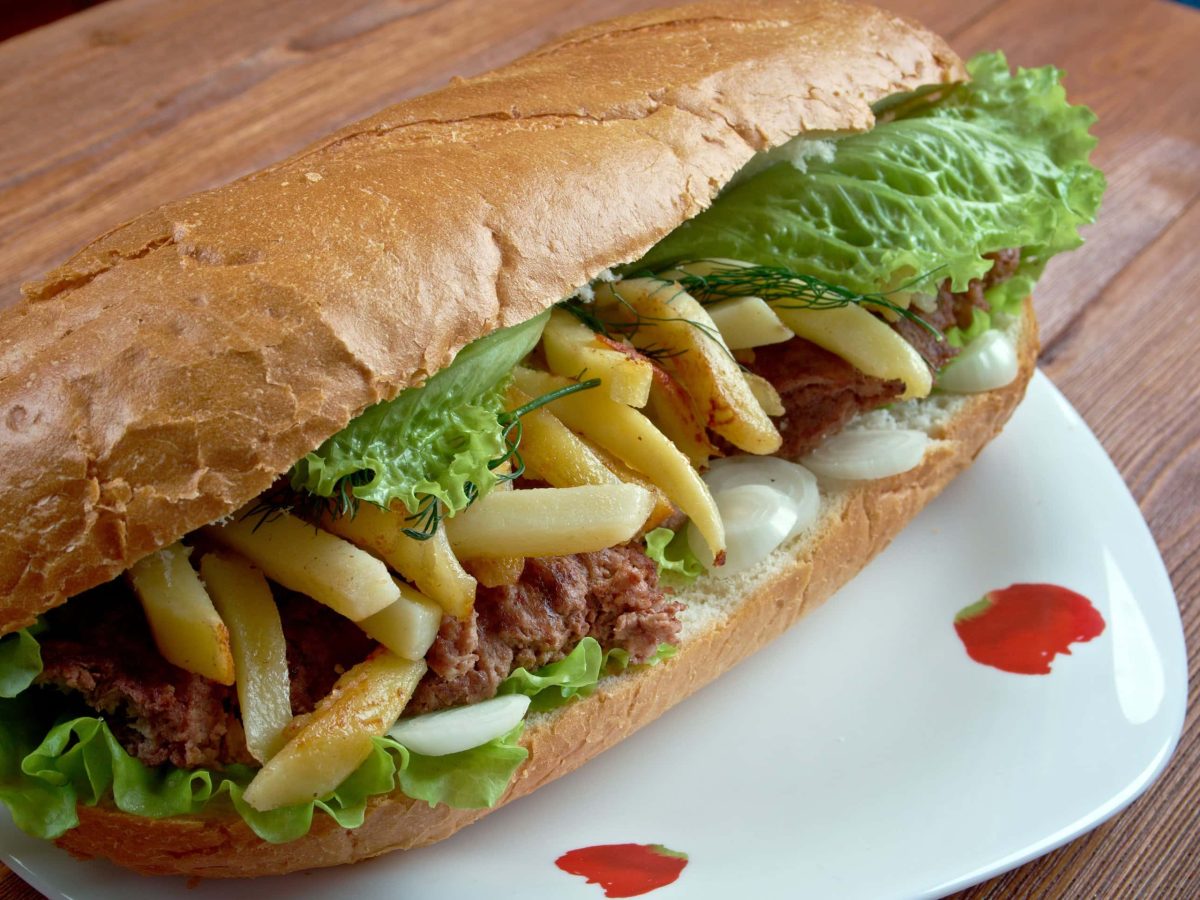 Streetfood Gatsby Sandwich mit Pommes und Masala-Steak auf weißem Teller und braunem Tisch. Frontalansicht.