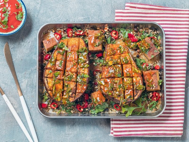 Ein silbernes Backblech mit gebackener Aubergine mit Harissa-Sauce auf einem rot-gestreiften Küchentuch