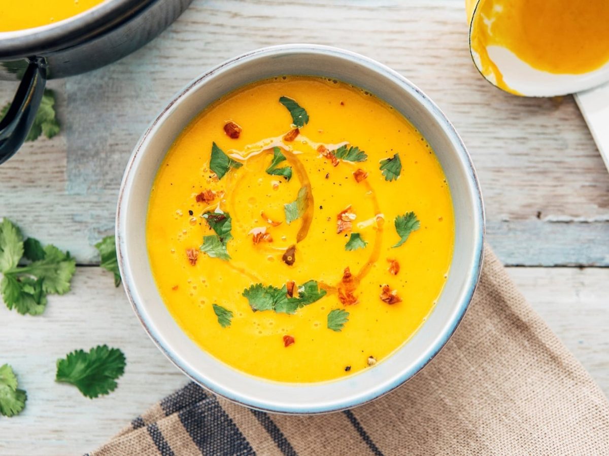 Eine Schüssel gelbe Gemüsesuppe mit Petersilie und Chiliflocken garniert, daneben eine Kelle, Draufsicht.