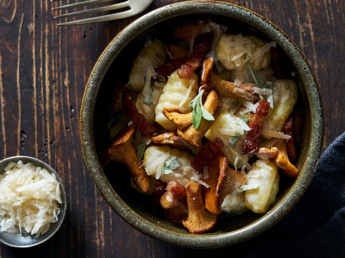 Eine graue Schüssel auf einem dunklen rustikalen Holztisch mit den Gnocchi und Pfifferlingen direkt von oben fotografiert. Daneben eine blaue Tischdecke und eine kleine Schale mit Parmesan auf der linken Seite.