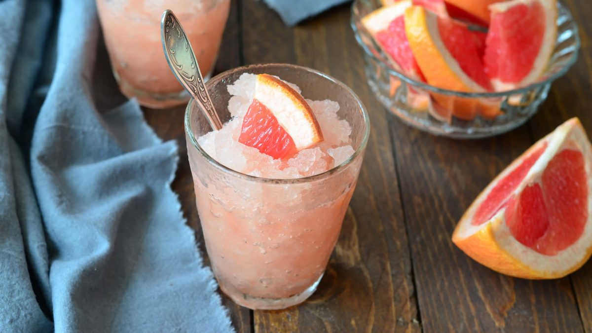 Ein Glas mit der Granita und einer angeschnittenen Grapefruit auf einem dunklen Holztisch mit einer blauen, gefalteten Tischdecke..