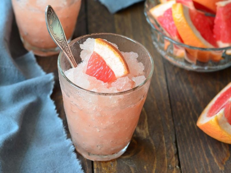 Ein Glas mit der Granita und einer angeschnittenen Grapefruit auf einem dunklen Holztisch mit einer blauen, gefalteten Tischdecke..