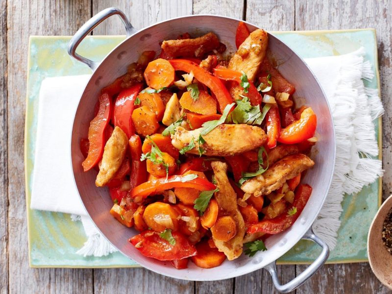 Hähnchen-Wok mit Paprika, Koriander und Möhren in der Pfanne vor hellem Hintergrund.