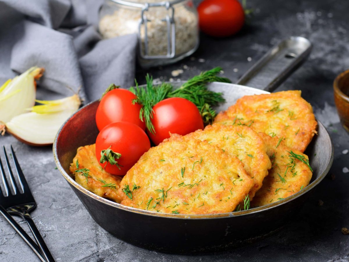 Ein dunkler tiefer Teller mit einigen Haferpuffern. Daneben ein paar Tomaten und eine angeschnittene Zwiebel auf einer dunklen Tischdecke.