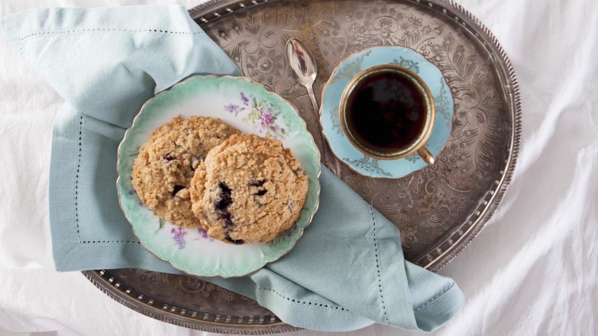 Zwei Heidelbeer-Streusel-Plätzchen auf einem ovalen Silbertablett und einer türkisenen Tasse mit Kaffee.