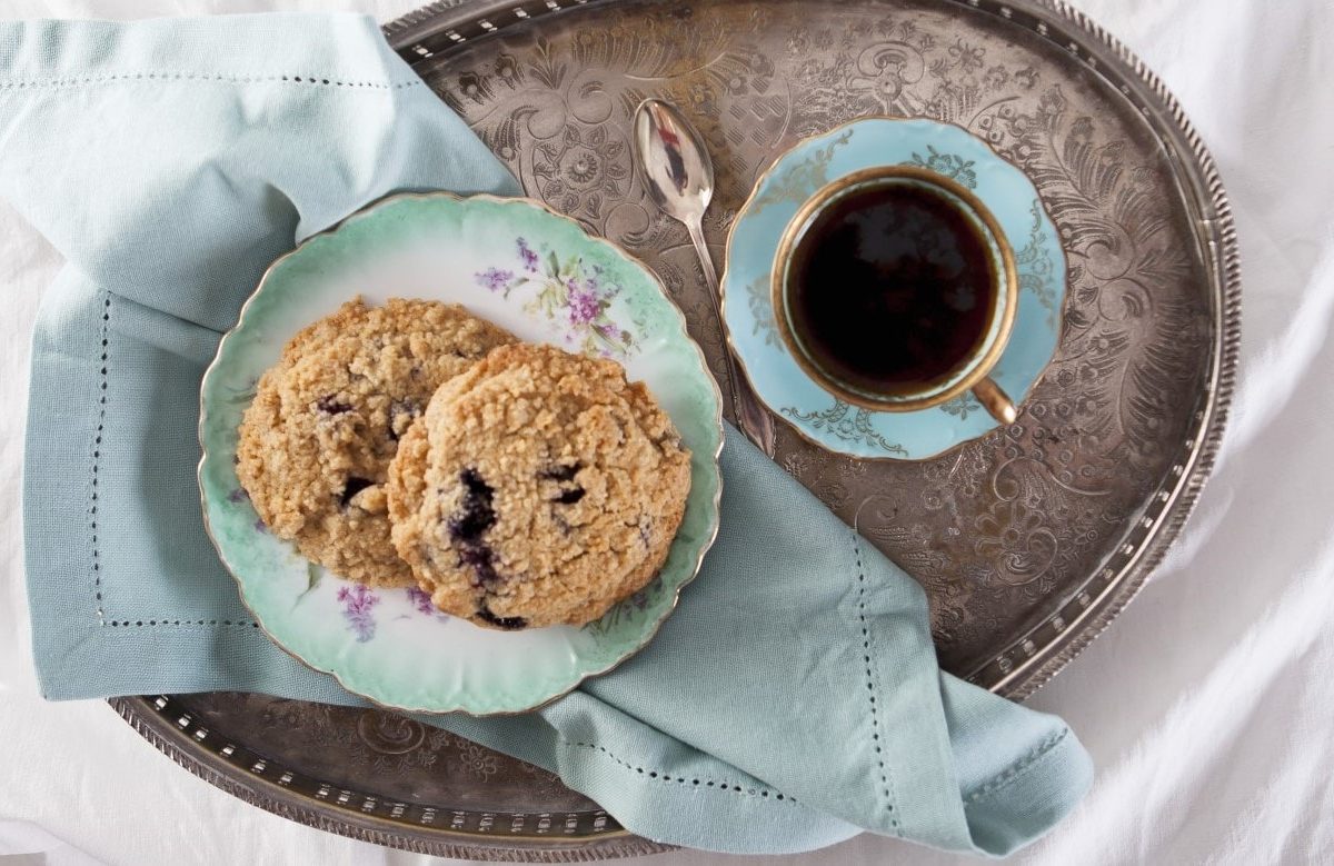 Zwei Heidelbeer-Streusel-Plätzchen auf einem ovalen Silbertablett und einer türkisenen Tasse mit Kaffee.