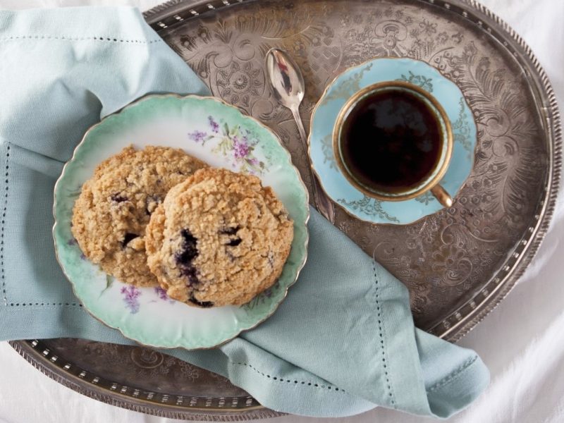 Zwei Heidelbeer-Streusel-Plätzchen auf einem ovalen Silbertablett und einer türkisenen Tasse mit Kaffee.