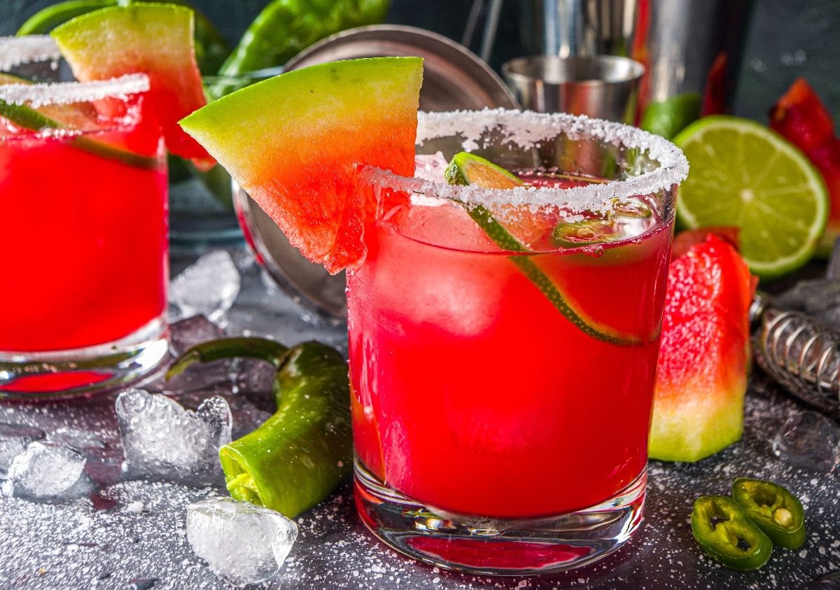 Mehrere rot-leuchtende Wassermelonen-Drinks auf einem grauen Untergrund mit Eis, Chili und Wassermelonenstückchen.