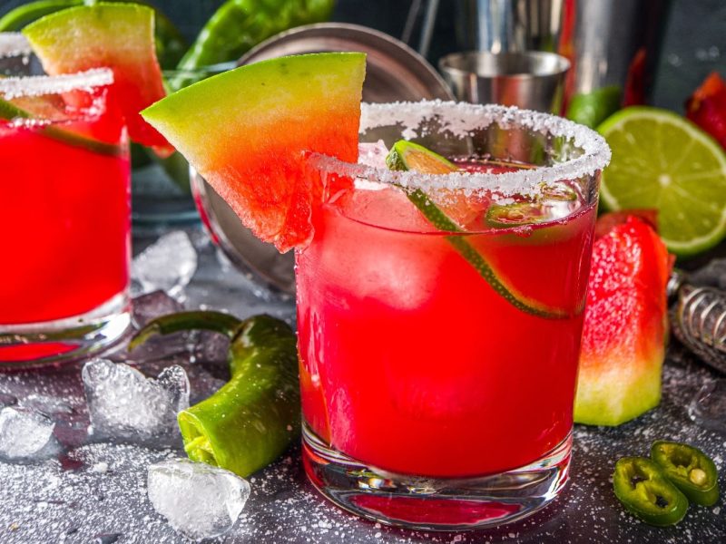 Mehrere rot-leuchtende Wassermelonen-Drinks auf einem grauen Untergrund mit Eis, Chili und Wassermelonenstückchen.