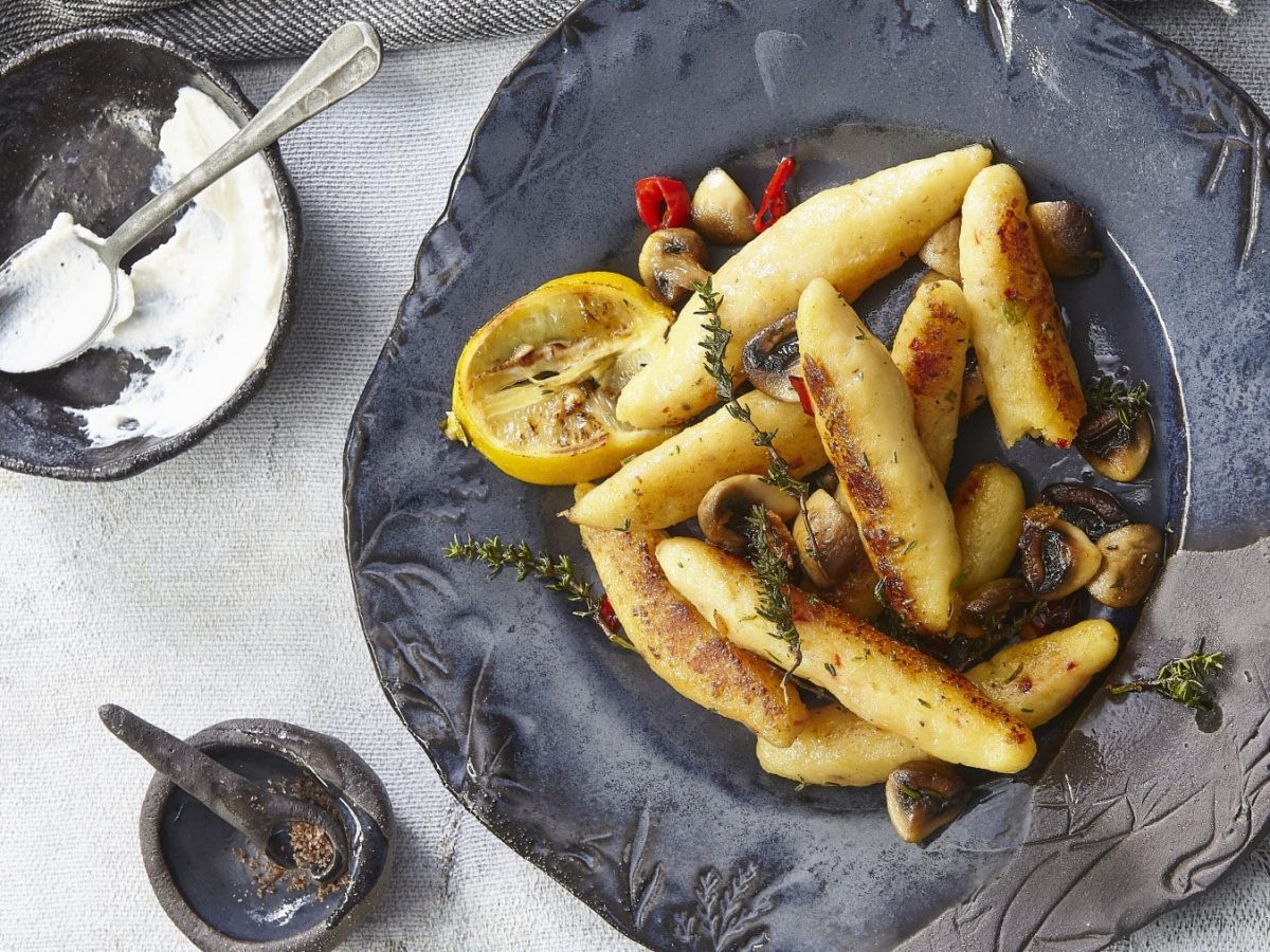 Kartoffelgnocchi mit Champignons und Sauerrahm garniert mit Zitrone, Thymian und Chili auf einem dunklen Teller neben einer Schale mit Sauerrahm vor grauem Hintergrund.