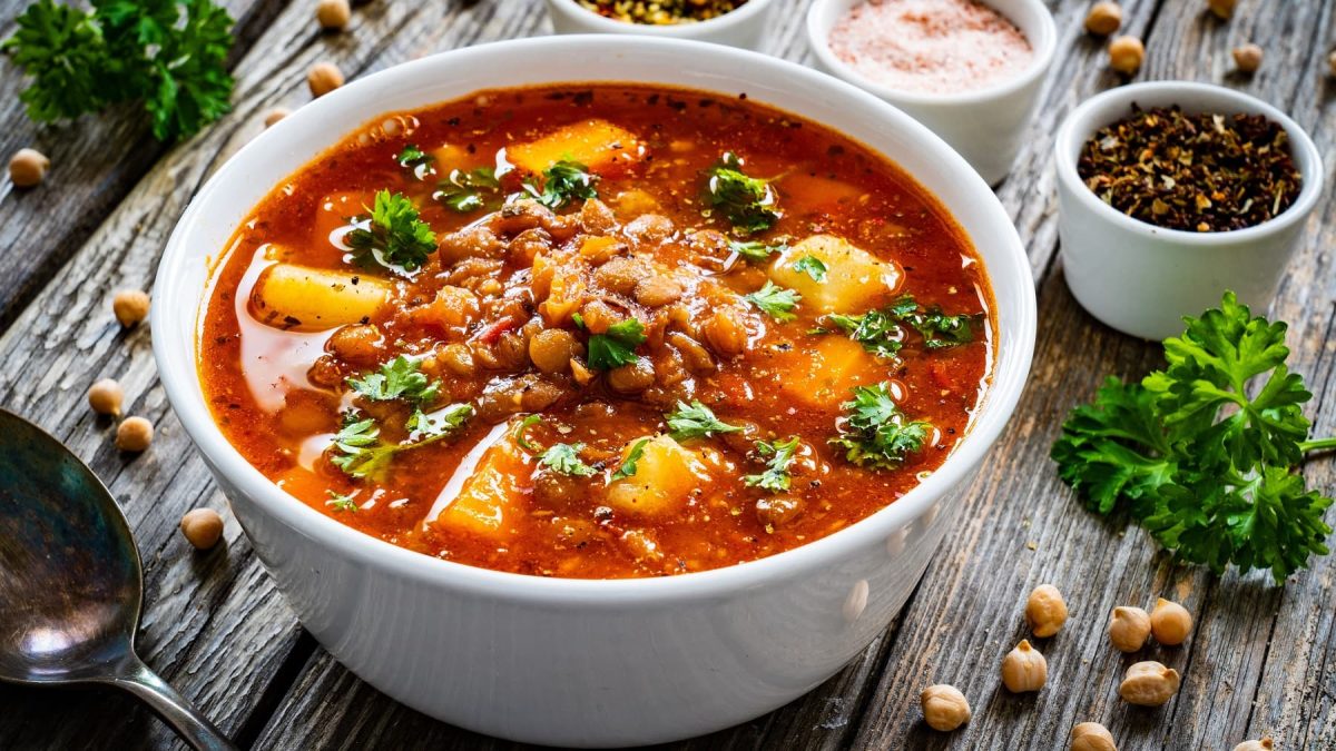 Eine weiße Schüssel Kichererbsensuppe mit Kartoffeln und frischer Petersilie, daneben stehen Schälchen mit Gewürzen und es liegen ein paar Kichererbsen herum.