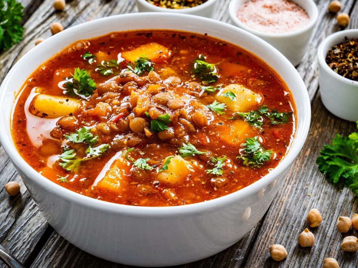 Eine weiße Schüssel Kichererbsensuppe mit Kartoffeln und frischer Petersilie, daneben stehen Schälchen mit Gewürzen und es liegen ein paar Kichererbsen herum.