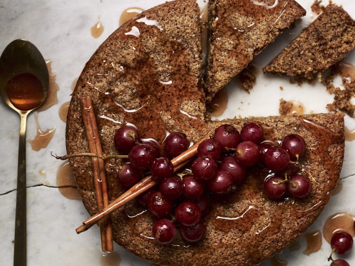 Runder Kokos-Haselnuss-Kuchen mit Trauben und Zimtstangen darauf neben einem Löffel mit Honig vor hellem Hintergrund