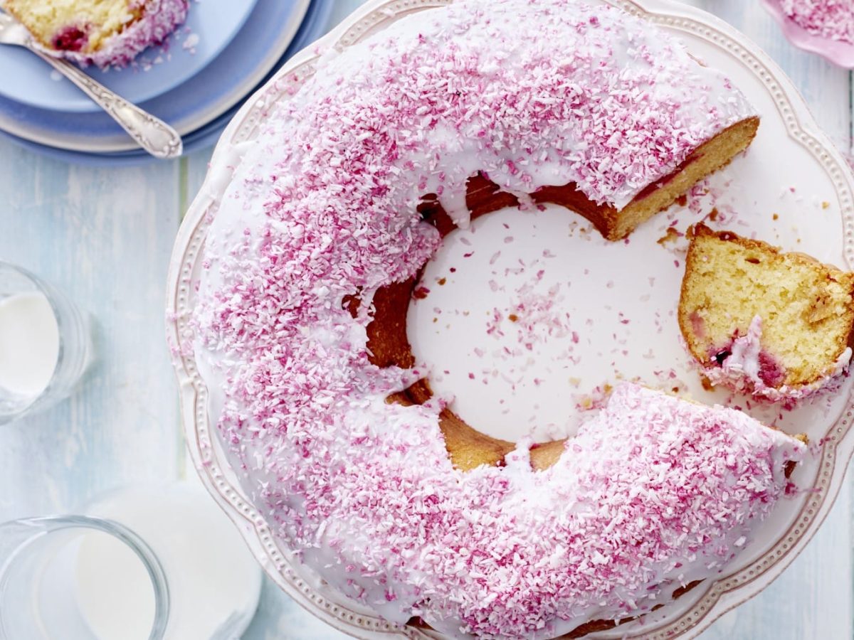 Runder Kokos-Himbeer-Kuchen mit Loch in der Mitte, weißer Glasur und rosa Raspeln darauf vor hellem Hintergrund neben Geschirr