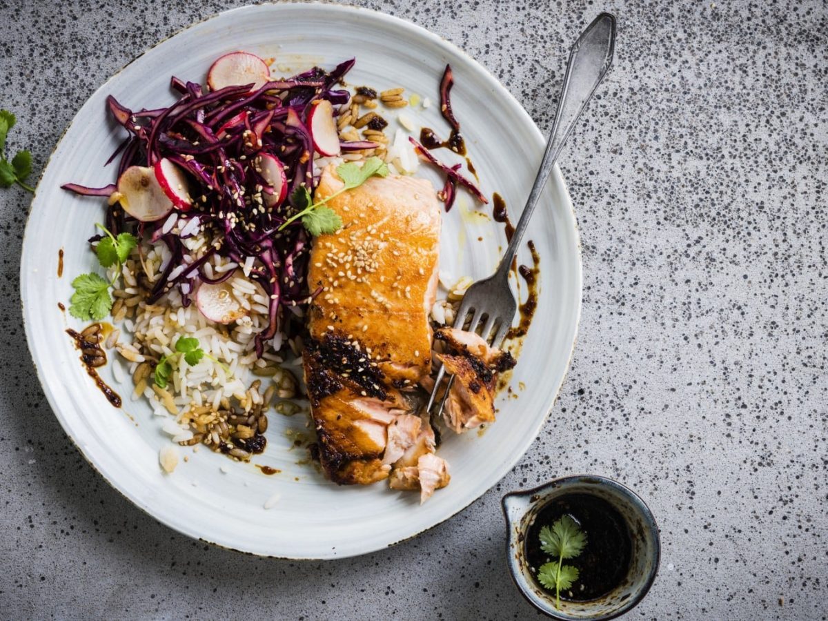 Lachs mit Rotkohl-Kimchi und Reis auf einem weißten Teller mit einer Gabel. Daneben steht ein Schälchen mit Sojasauce.