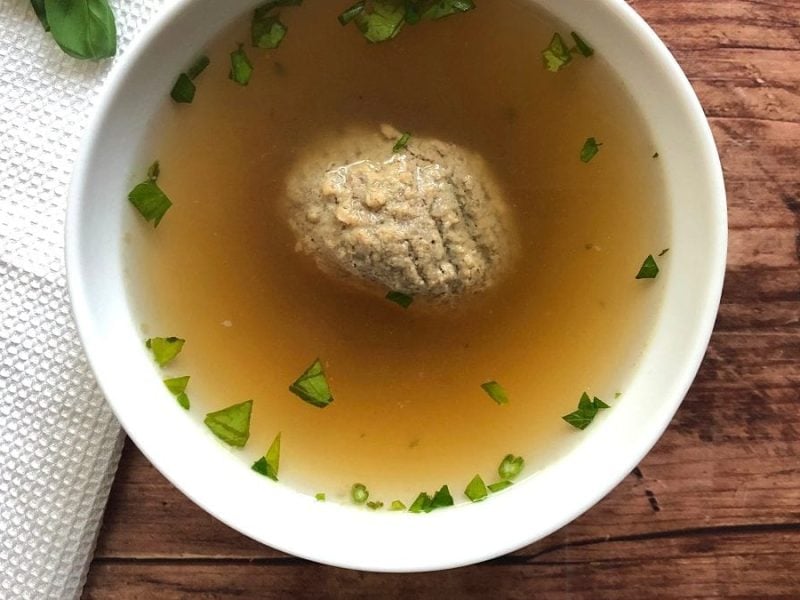 Aus der Vogelperspektive: Eine Schüssel Leberknödelsuppe. Daneben liegen Kräuter.