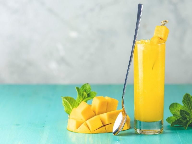 Ein Glas Mango-Limonade mit Ginger Beer und frischen Mangostücken, daneben liegen Minzblätter und eine aufgeschnittene Mango.