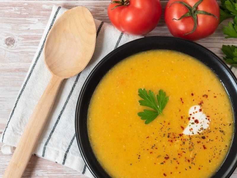 Aus der Vogelperspektive: Eine Schüssel Mercimek çorbasi steht auf einem Tisch. Es handelt sich um eine rote Linsenduppe. Daneben liegen Tomaten und frische Petersilie.