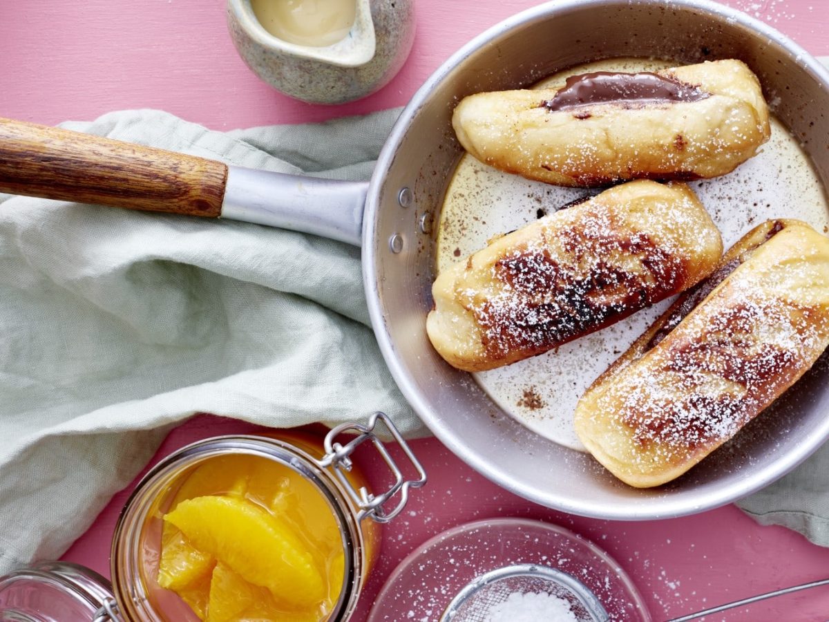 In einer Pfanne sind drei gefüllte Milchbrötchen mit Orangenkompott in einem Glas daneben vor rosa Hintergrund. Die Milchbrötchen sind gefüllt mit Nuss-Nougat-Creme und bestäubt mit Puderzucker.