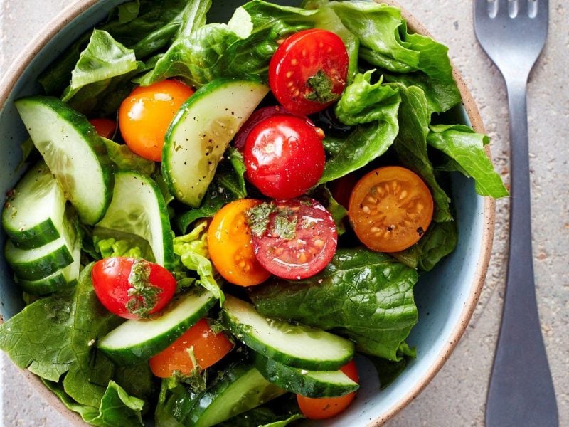 Schüssel mit grünem Salat, Tomaten und Gurkenscheiben und Minzdressing neben einer Gabel vor hellem Hintergrund.