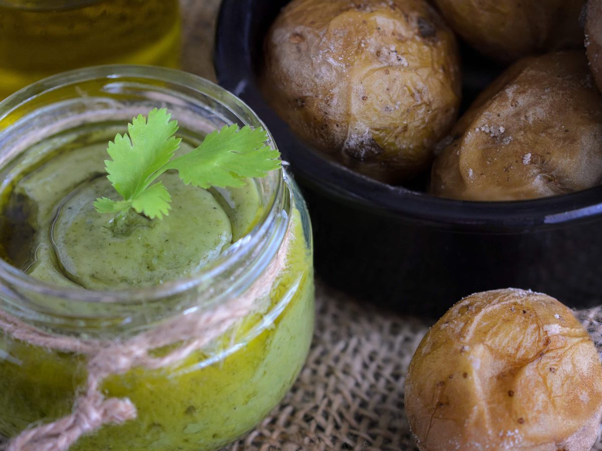 Mojo verde mit Paprika im Einmachglas auf hellem Untergrund. Daneben Patatas bravas und Olivenöl. Draufsicht.