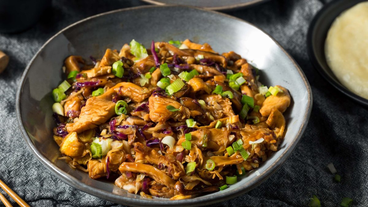 Eine gusseiserne Pfanne mit dem Mu Shu Pork auf einem grauen Tisch. Rechts oben am rand sieht man ein kleines Stück eines Pfannkuchens.