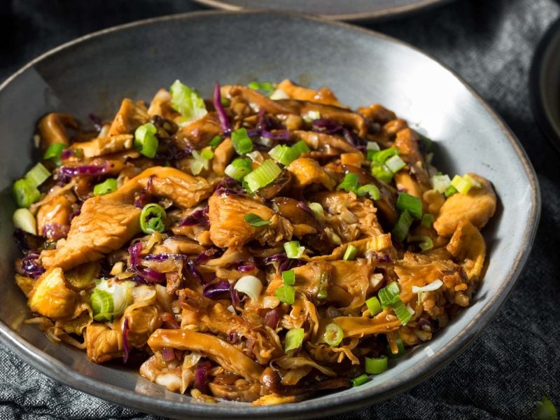 Eine gusseiserne Pfanne mit dem Mu Shu Pork auf einem grauen Tisch. Rechts oben am rand sieht man ein kleines Stück eines Pfannkuchens.